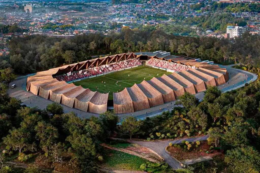 O que há por trás do primeiro estádio 100% em madeira do Japão? 5 a imagem mostra como será o primeiro estádio 100% em madeira do japão. O estádio tem uma cobertura em madeira e está cercado por árvores.