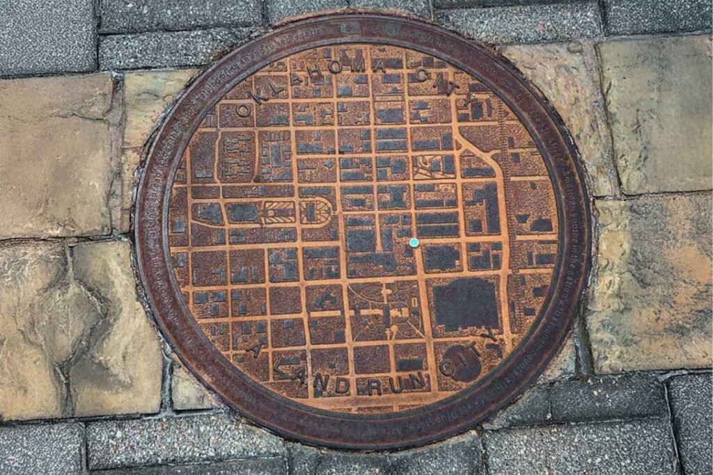 Porque tem um mapa nas tampas de bueiro de Oklahoma City? 3 As tampas de bueiro de Oklahoma City possuem um mapa da cidade. A imagem mostra um exemplo disso com uma tampa de bueiro com o mapa e um ponto indicando a localização da peça no mapa.